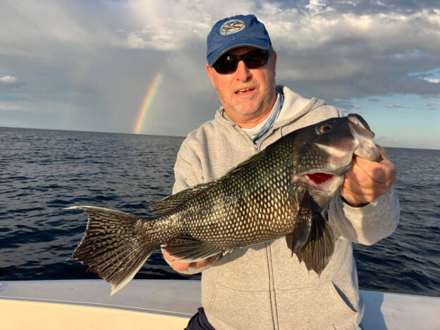 A man on a boat proudly holding a large fish against the backdrop of the open ocean. He’s wearing a gray hoodie, blue cap, and sunglasses. A vivid rainbow appears in the cloudy sky behind him, adding a striking, serene element to the seascape.