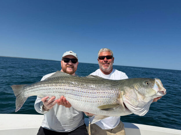 Two smiling men on a boat holding up a large striped bass with the ocean and horizon behind them.